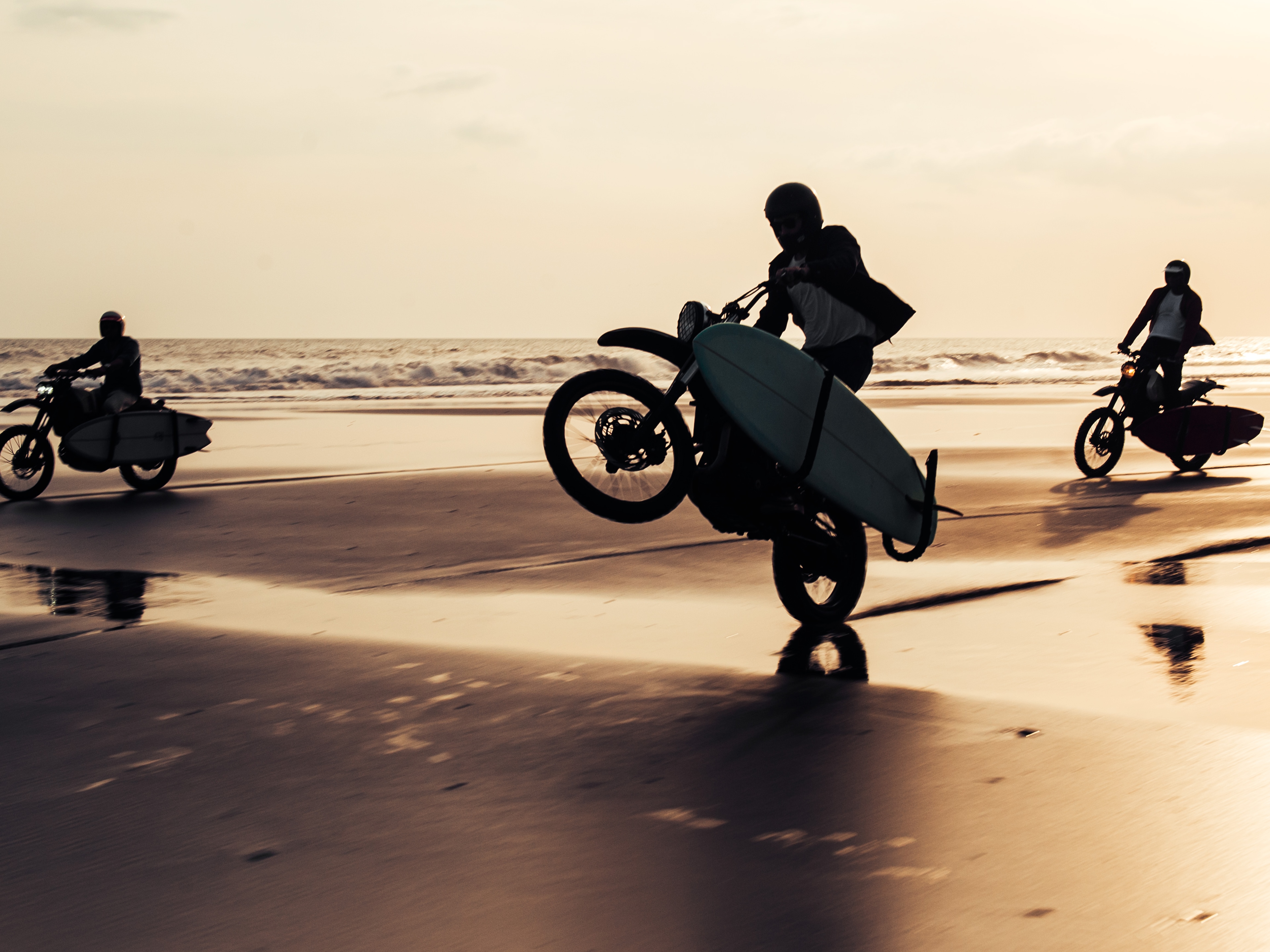 日落時分，三輛拖著衝浪板的 Deus Ex Machina 摩托車在海灘上馳騁。